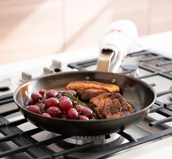 A Misen Pre-Seasoned Carbon Steel pan on a gas cooktop with seared pork chops, fresh thyme, and roasted red grapes — a restaurant-quality dinner mid-finish.
